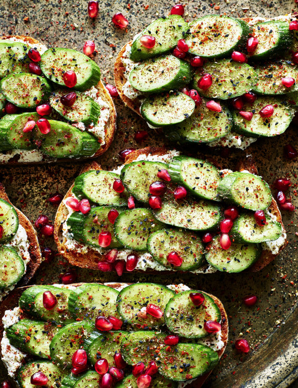 Food photography showing thick slices of bread, smothered in feta cheese, cucumber slices and pomegranate seeds