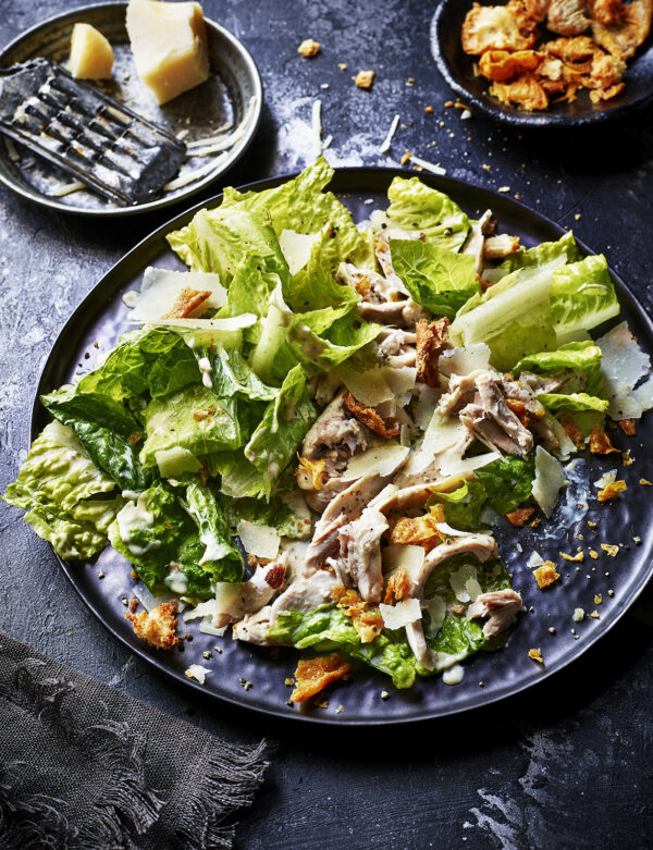 Food product photography showing a plate of chicken caesar salad with some extra croutons and parmesan cheese on the side.